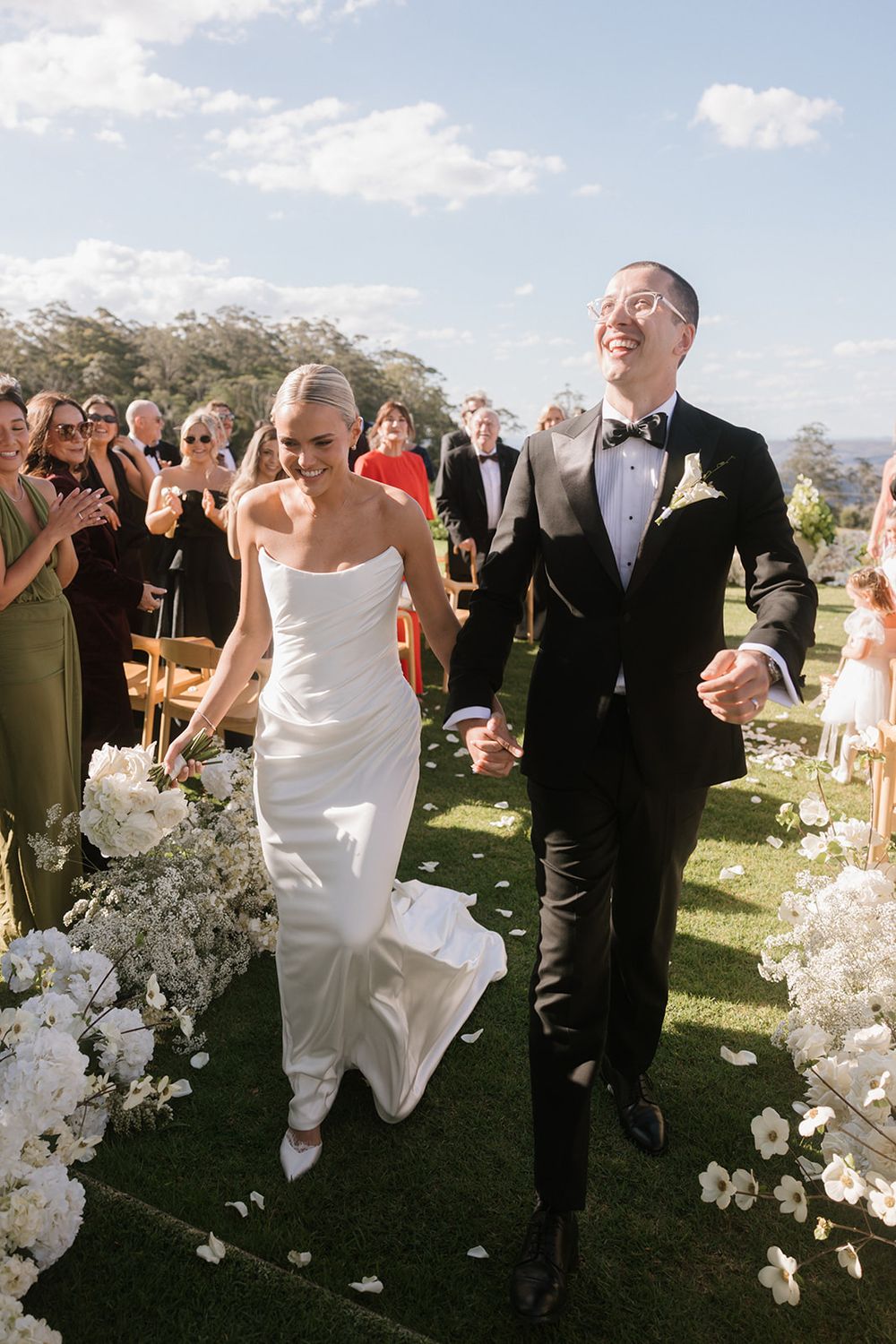 Bride and groom walk joyfully down outdoor aisle lined with white flowers after wedding ceremony.