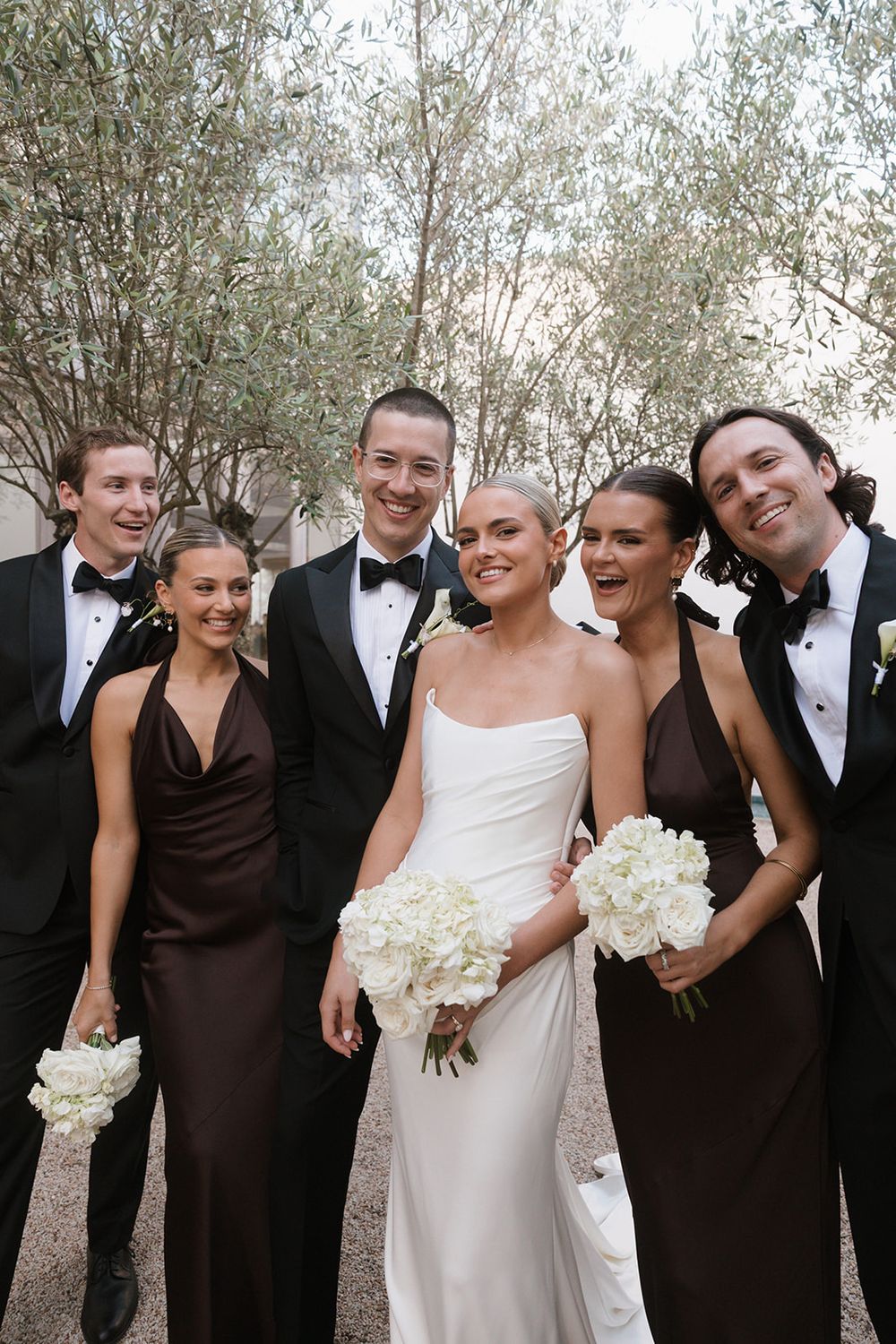 Wedding party poses outdoors, bride in white gown with bouquet, bridesmaids in brown dresses, groomsmen in black tuxedos.