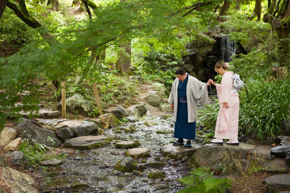 Two people in traditional Japanese kimono walk along a rocky stream path in a lush green garden.