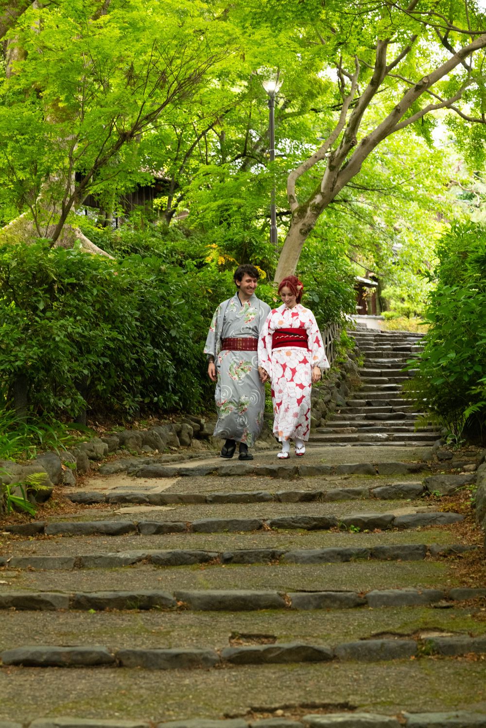 Couple wearing traditional Japanese kimonos walking down stone steps in a lush green garden.