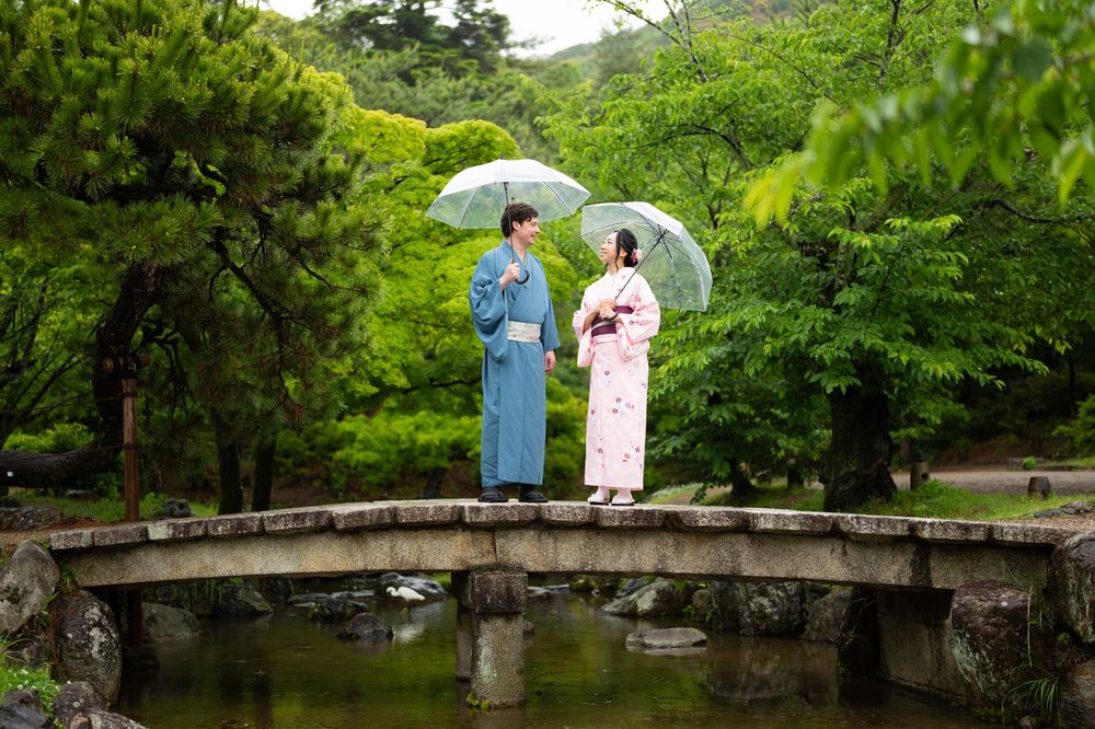 Two people in traditional Japanese kimonos holding umbrellas on a stone bridge surrounded by lush green gardens.