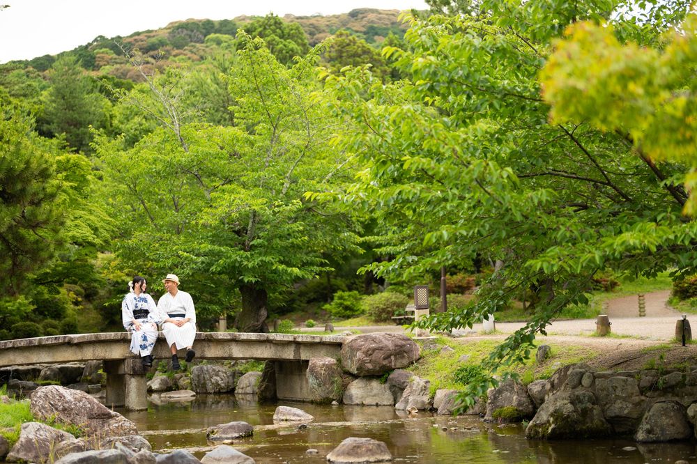 Two women in traditional kimonos sit on a stone bridge in a lush Japanese garden with a serene pond.