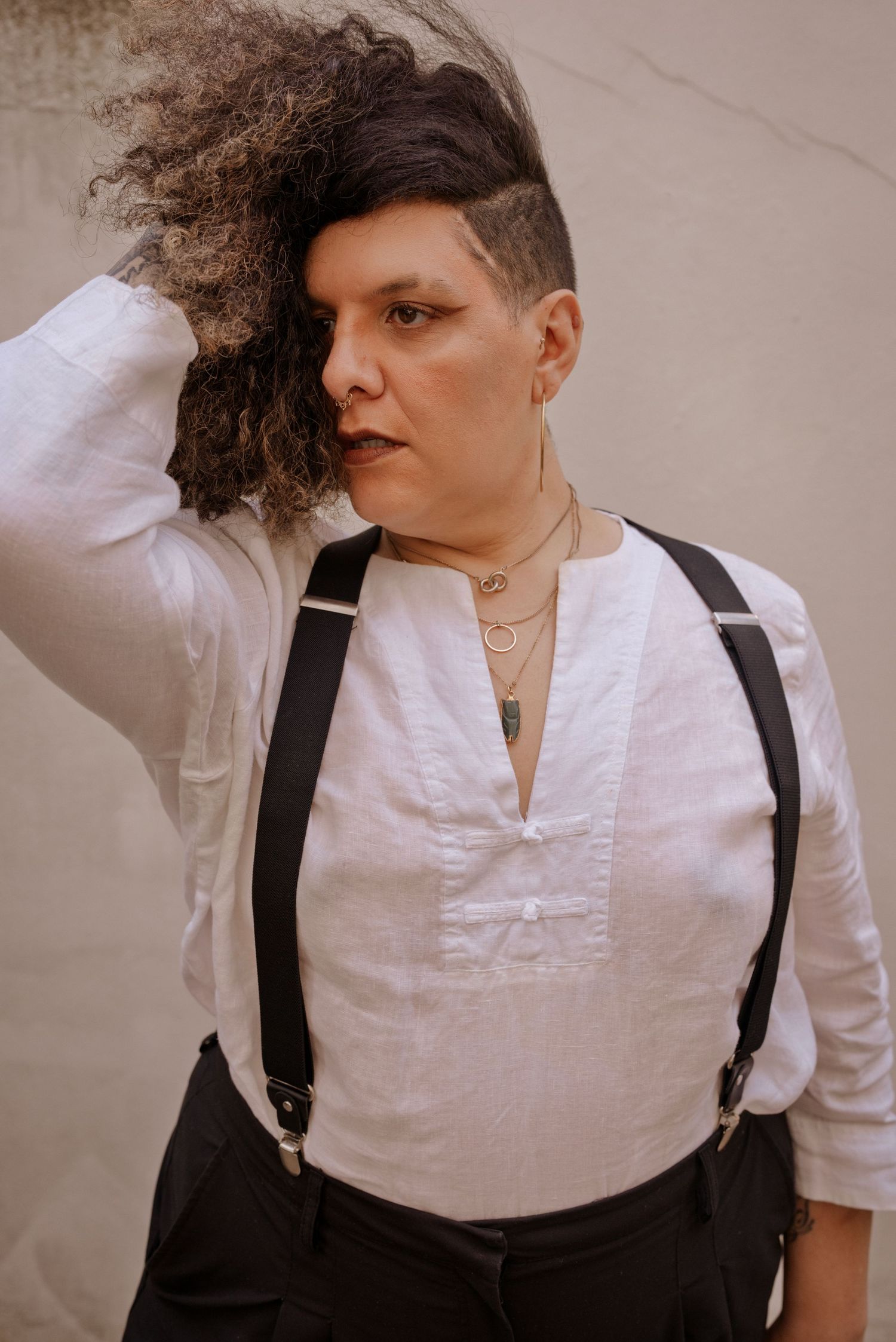 Person with curly undercut hair wearing white linen shirt with suspenders and layered necklaces poses against wall.