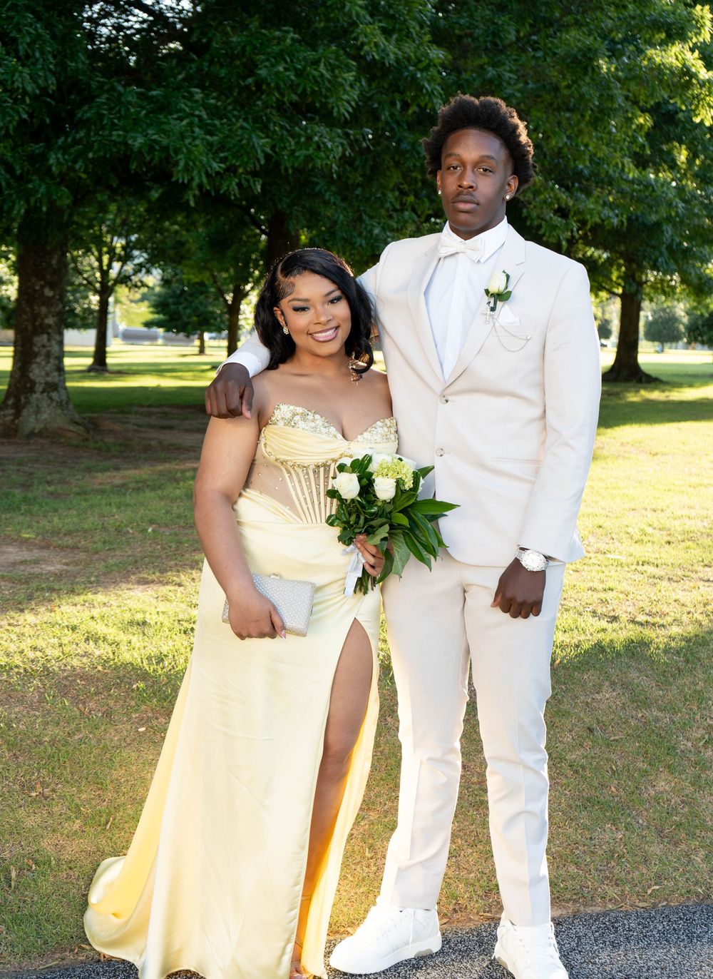 A couple dressed in prom attire, woman in yellow gown and man in white suit, posing outdoors in a park.