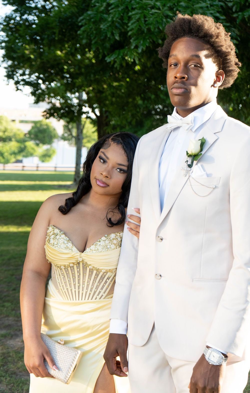 A couple dressed in formal prom attire, woman in yellow gown and man in white tuxedo, posing outdoors.