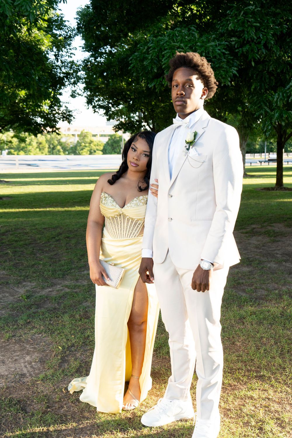 A young couple dressed in prom attire, woman in yellow gown and man in white suit, posing outdoors in a park.