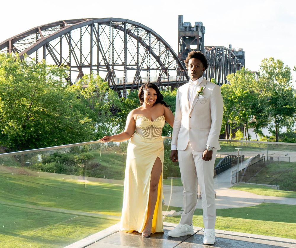 A couple in formal attire poses before a steel bridge, woman in yellow gown, man in white suit.