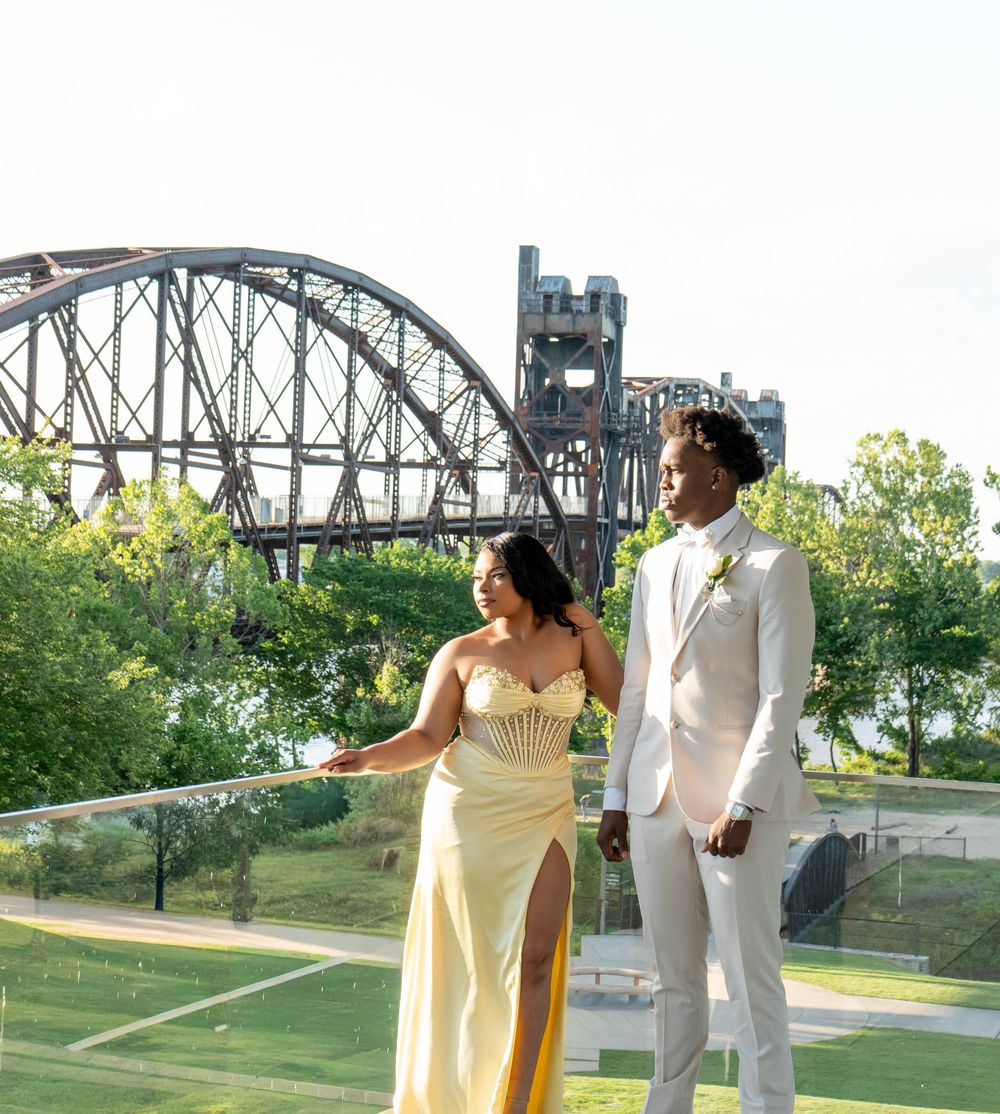 Couple in formal attire posing on a rooftop with a steel arch bridge and green trees in the background.