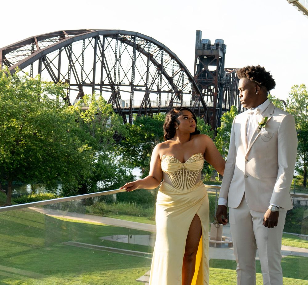 Couple in formal attire posing outdoors near a steel bridge, woman in gold gown, man in white suit.