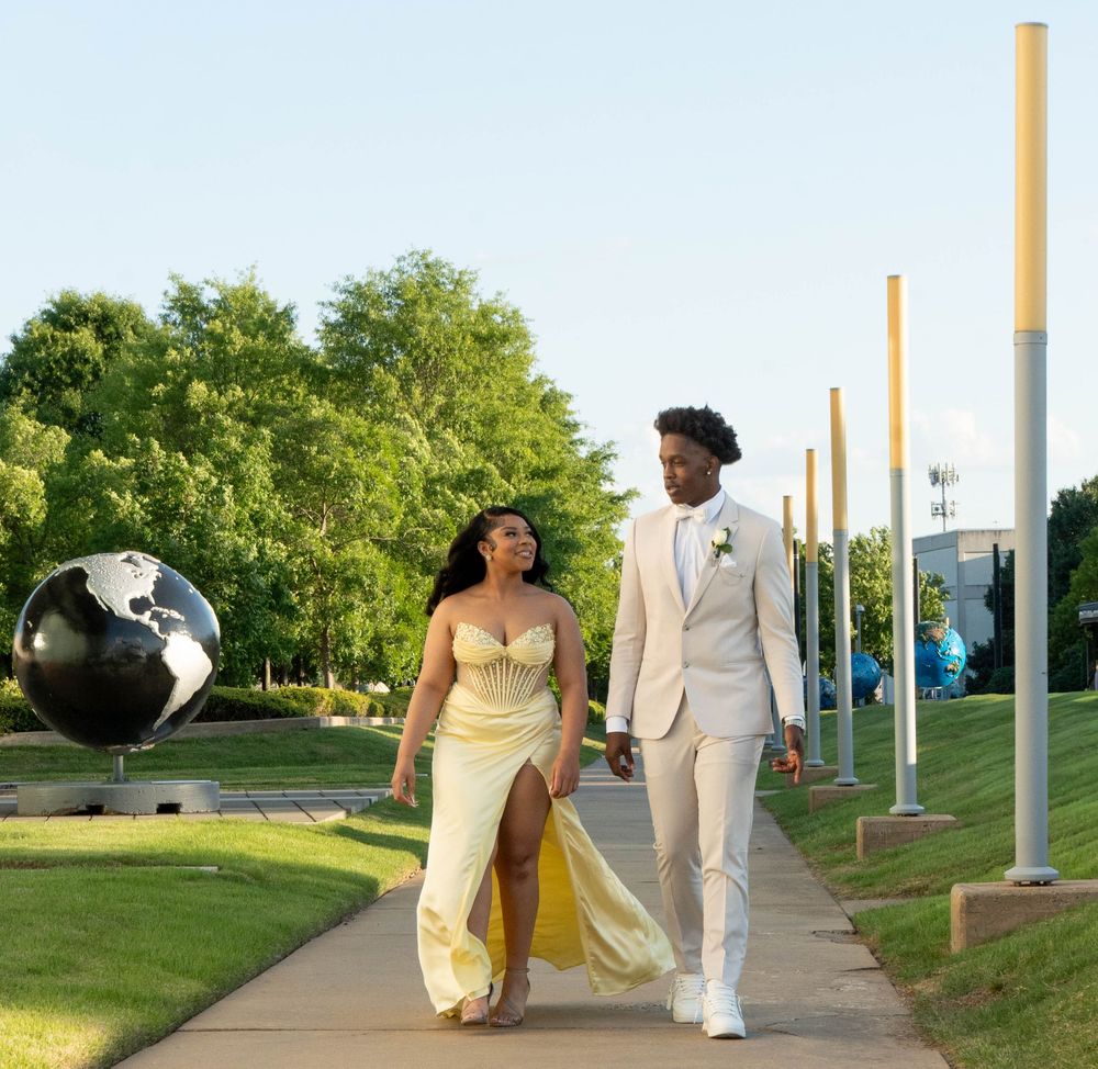 A couple dressed in formal attire walks along a garden path, wearing a yellow gown and white suit for prom.