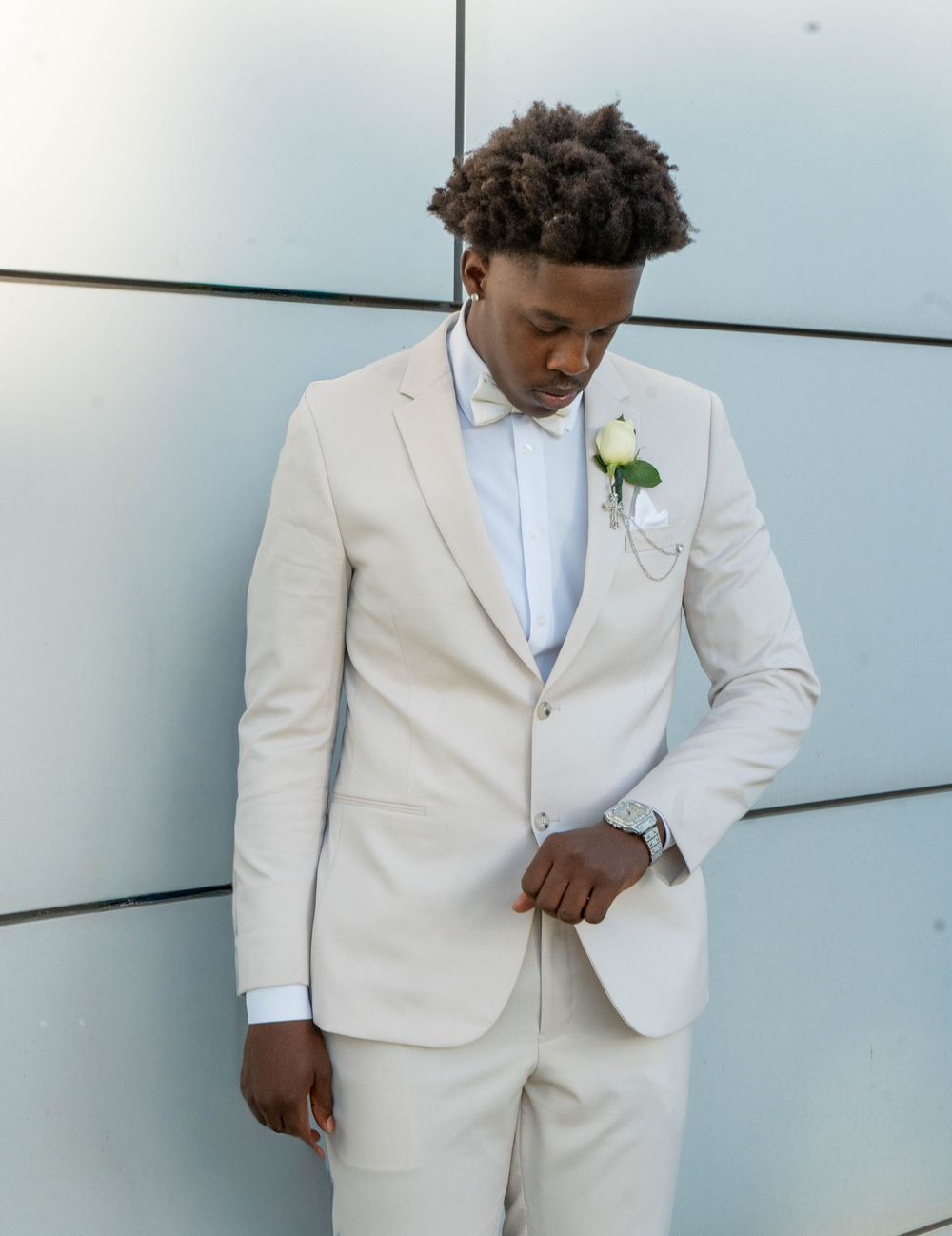 A young Black man in a cream suit with boutonniere and bow tie looks down elegantly against a modern wall.