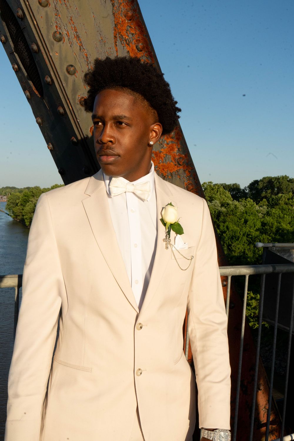 Young man in cream tuxedo with white boutonniere poses on bridge overlooking river at sunset.