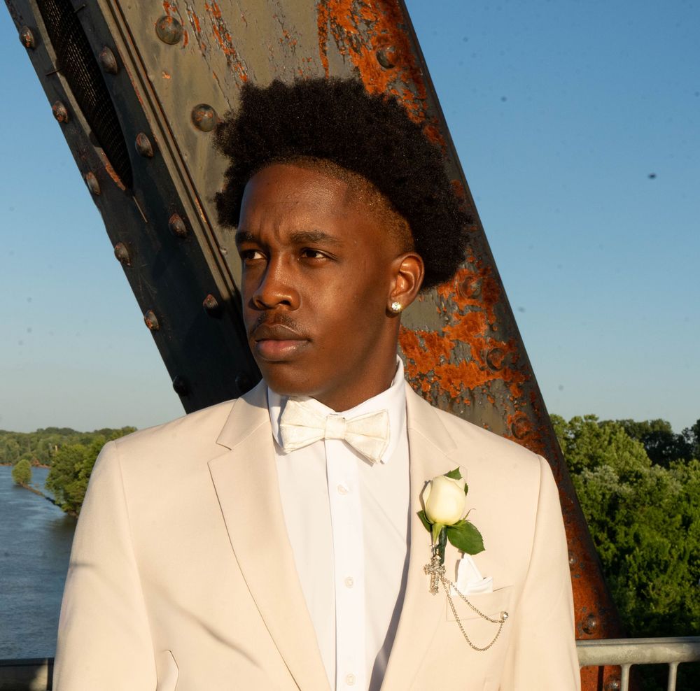 Young man in white tuxedo with boutonniere and bow tie posing outdoors near water at dusk.