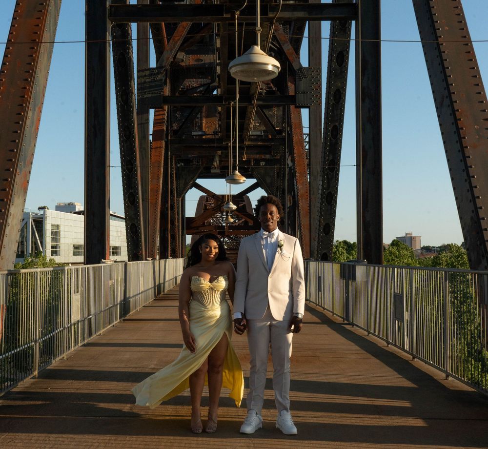 Couple in formal attire posing on a historic iron bridge at golden hour, woman in yellow gown, man in white suit.