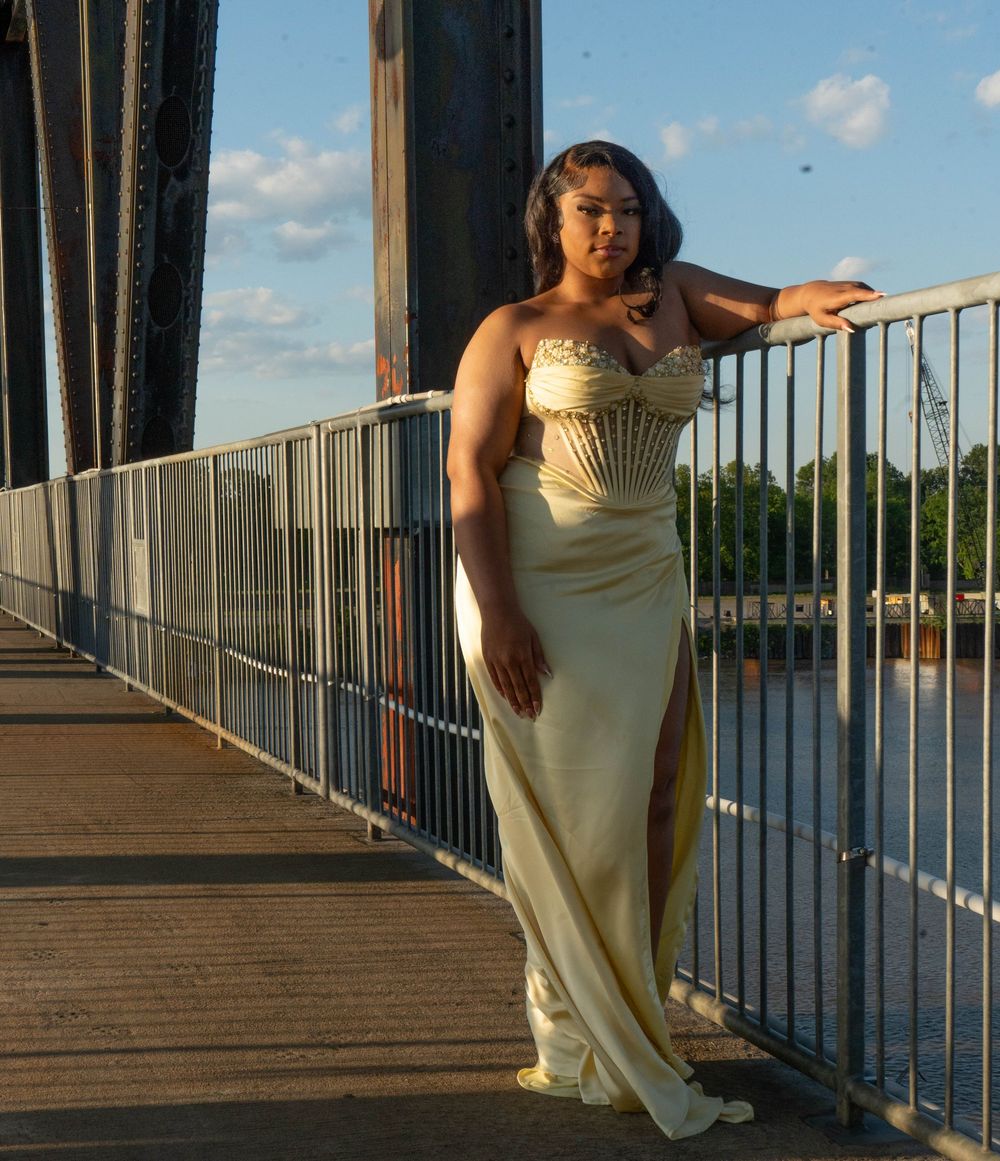 Woman in gold strapless gown posing on bridge railing at sunset with river in background.