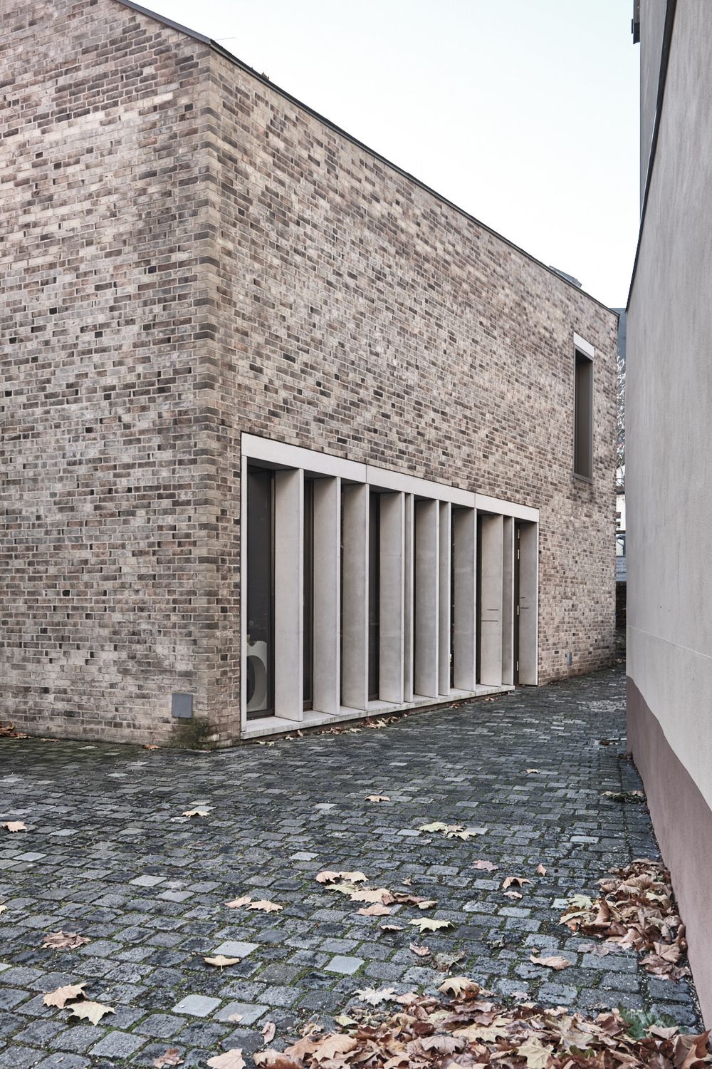 Modern brick building with tall vertical concrete-framed windows beside a cobblestone alley covered in fallen autumn leaves.