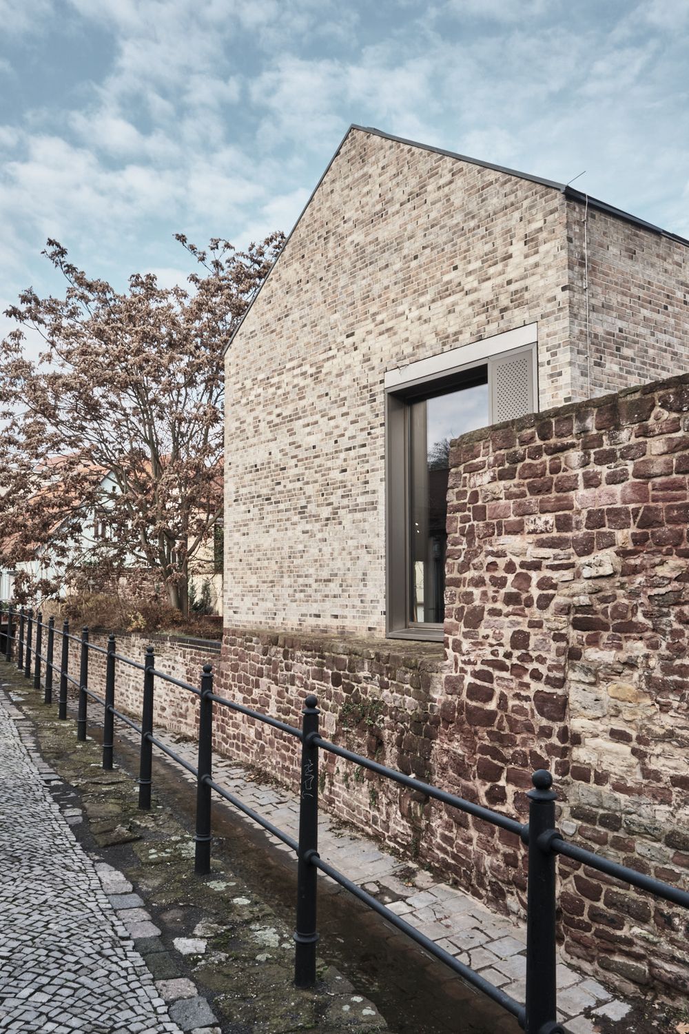 Modern brick house with a gabled roof and recessed window beside an old red sandstone wall, black railing and cobblestone path.
