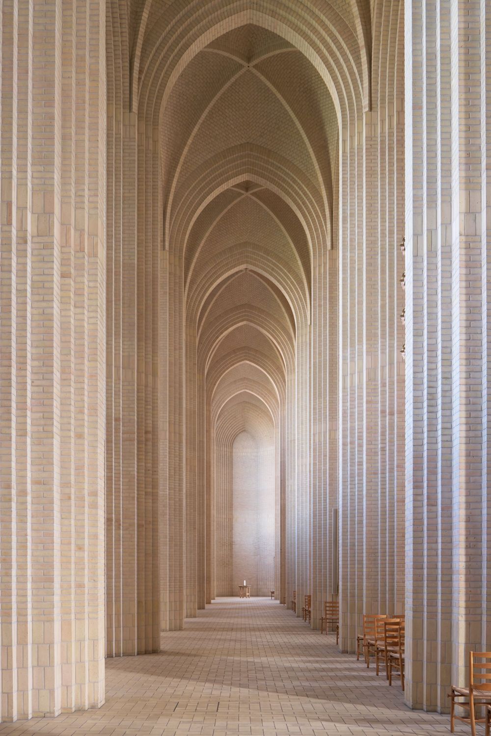 Tall brick pillars and layered pointed arches line the long nave of Grundtvig's Church, with wooden chairs along the paved floor.