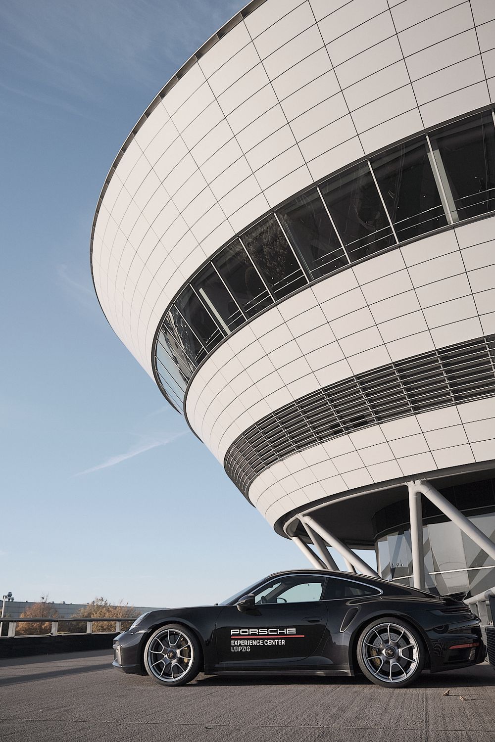 Black Porsche 911 parked beside the curved white Porsche Experience Center Leipzig building under a clear blue sky.