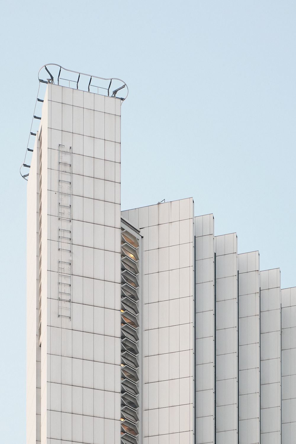 White modern high-rise building with stepped facade, exterior ladder and rooftop railing against a clear pale blue sky.