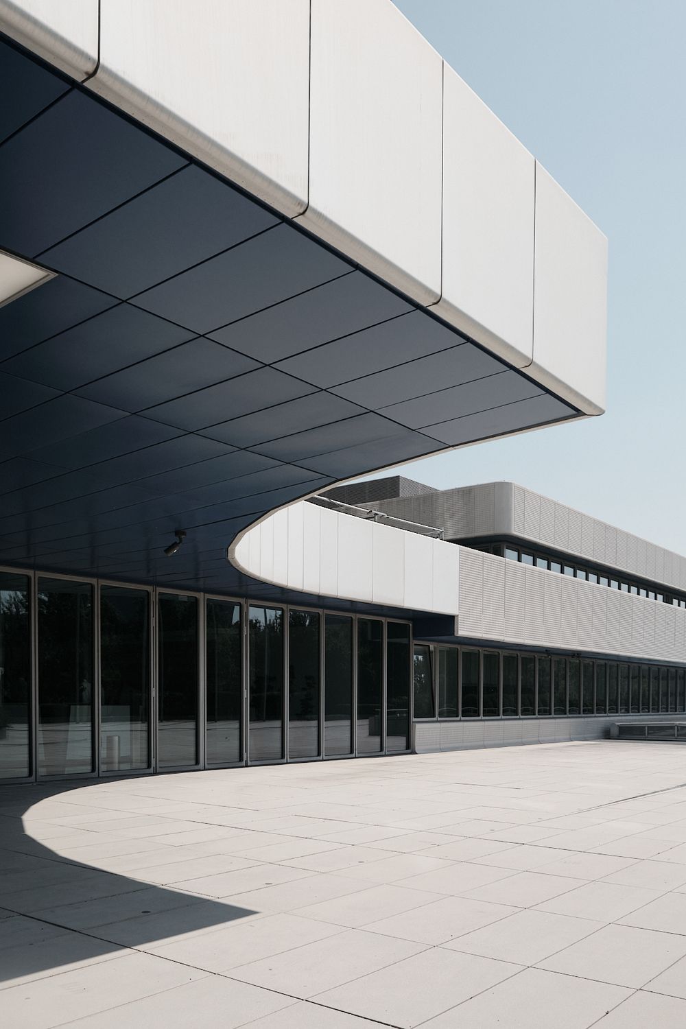 Modern building with a curved dark overhanging roof, white metal cladding and floor-to-ceiling glass windows beside a sunlit paved plaza.