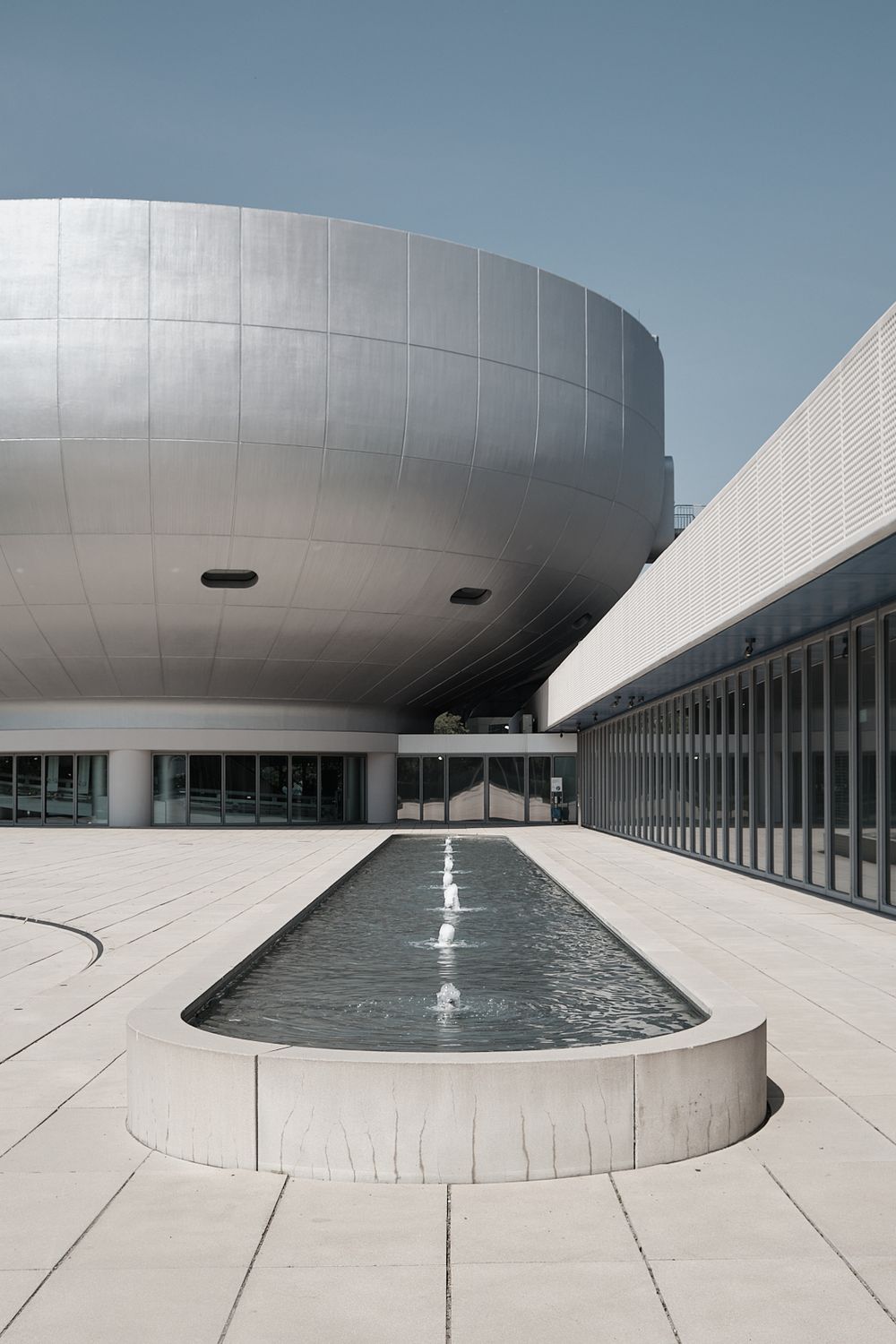 Modern museum plaza with a silver bowl-shaped building and a long rectangular reflecting pool with fountains beside a glass-walled wing.