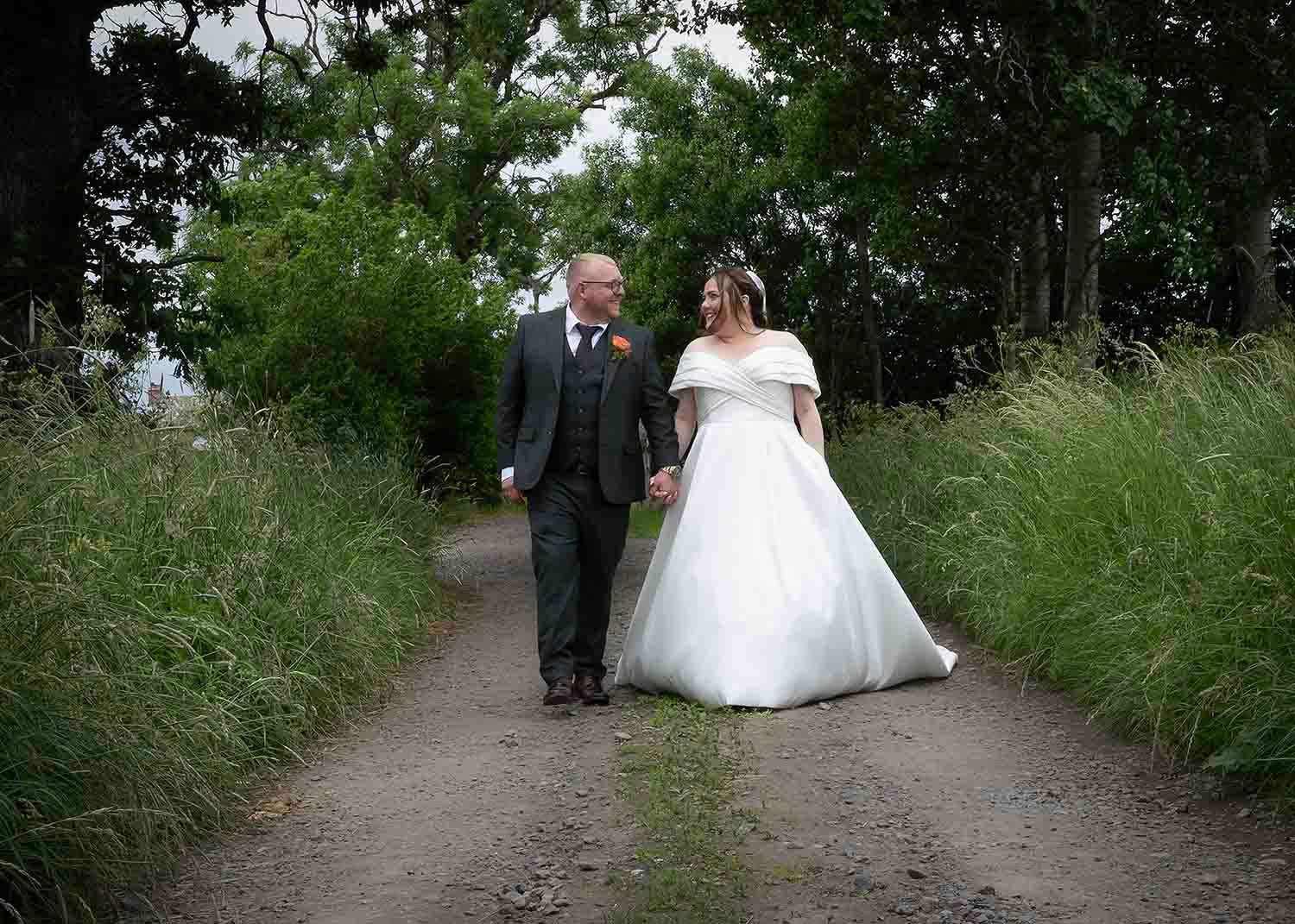 Lucas and Ellie walking hand in hand along a gravel path bordered by tall grass and trees at The Den at Four Acre.