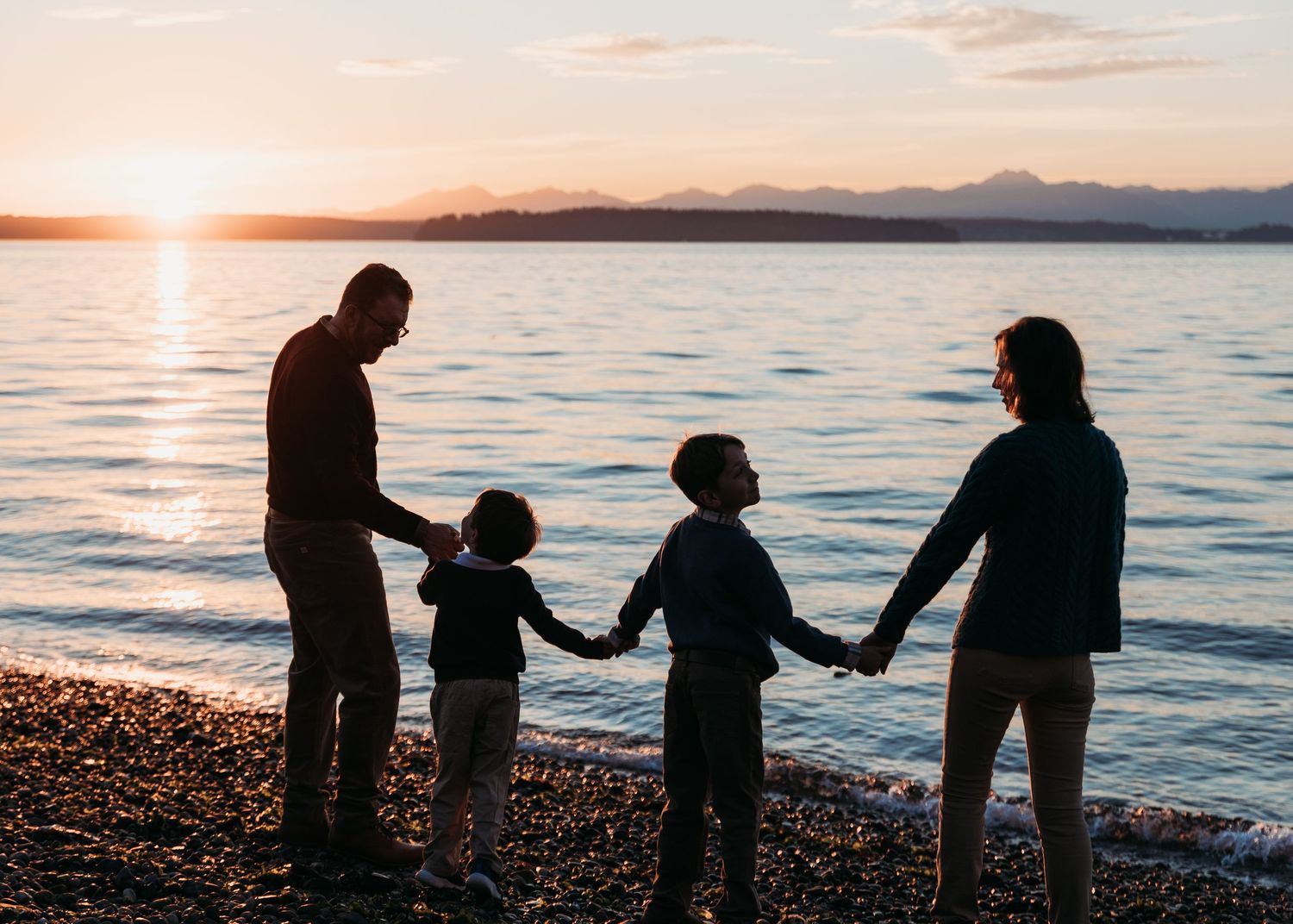 Silhouetted family of four holding hands on a pebble beach at sunset, with calm water and distant mountains in the background.
