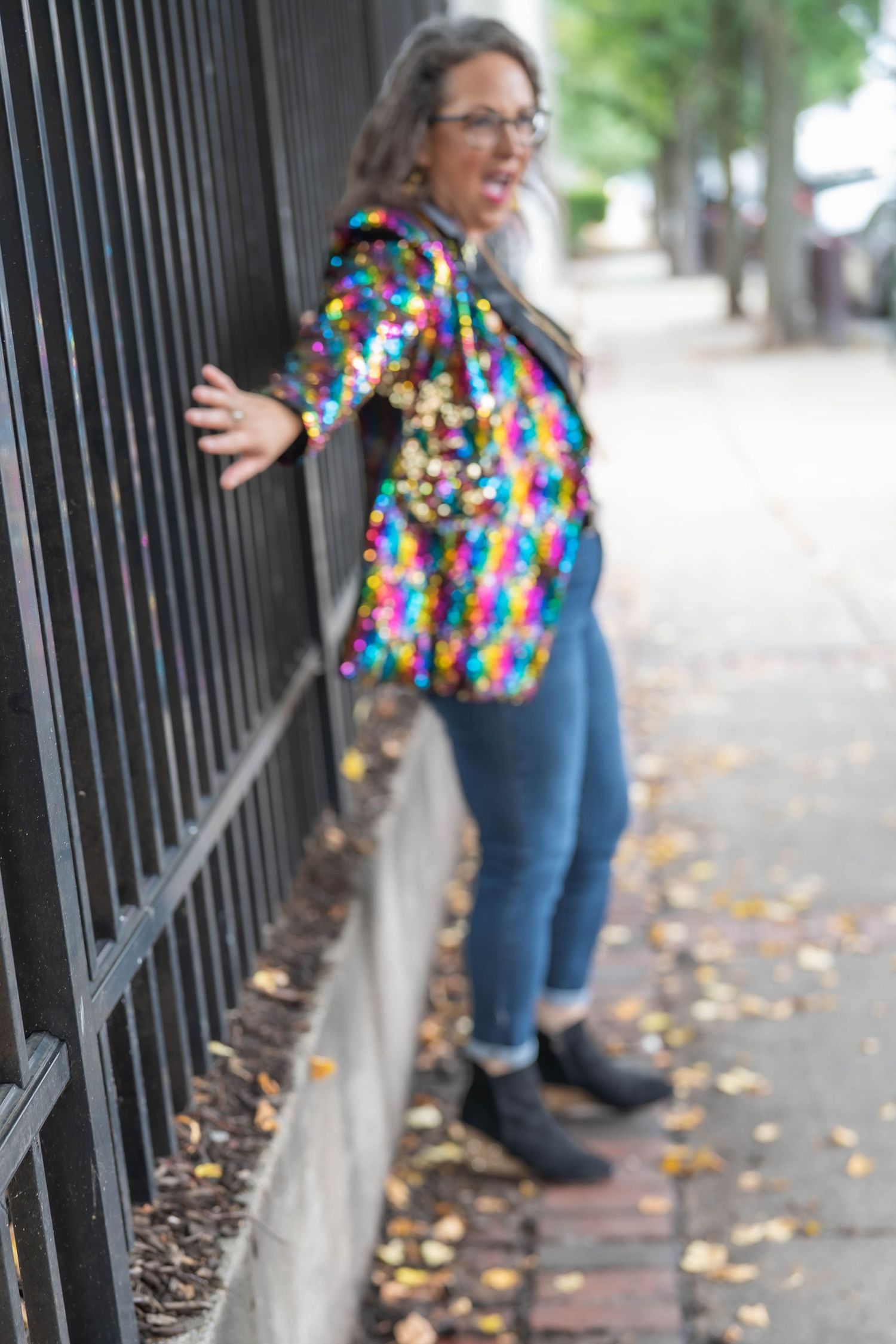 Woman in glasses and a colorful sequin jacket leans against a black iron fence on a brick sidewalk covered in fallen autumn leaves.