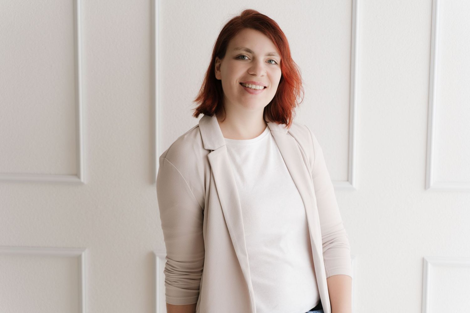Smiling redhead woman in beige blazer and white top posing confidently against a white paneled wall.