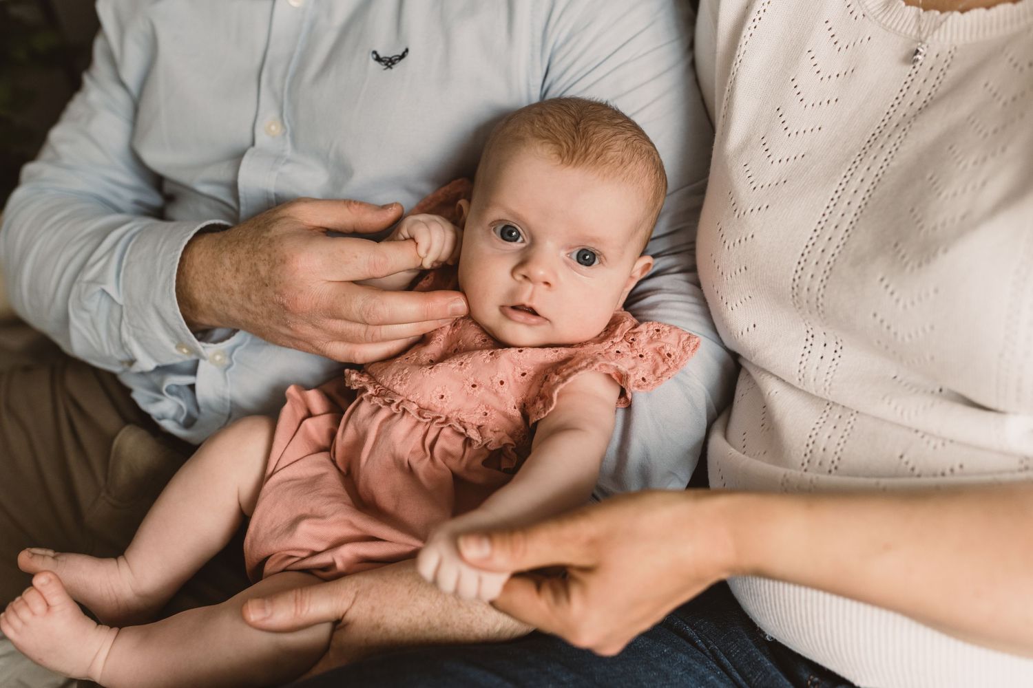 Newborn baby in pink romper cradled by parents hands in a soft, intimate family portrait session.