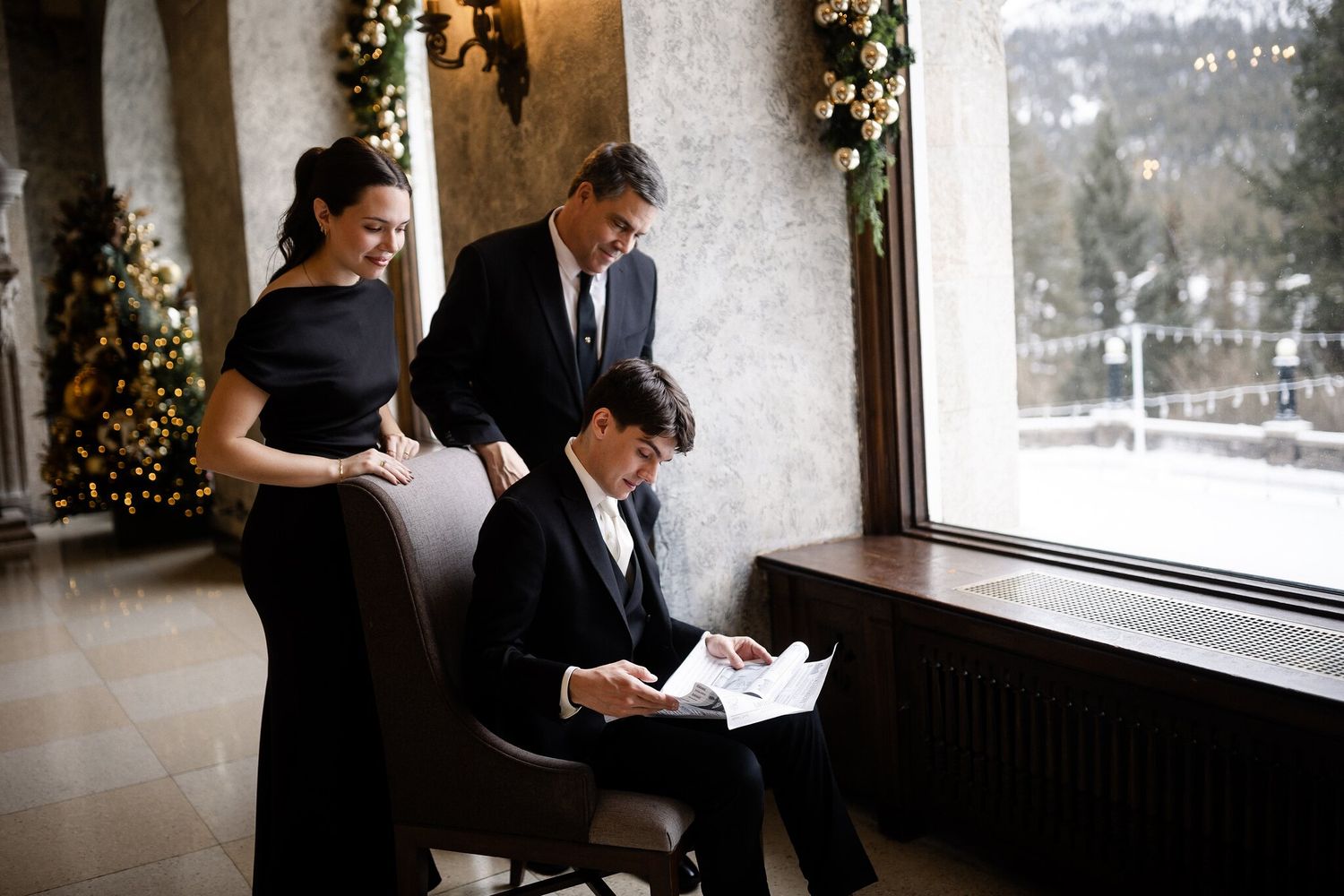 Groom reading by a castle window as two loved ones look on beside a Christmas tree at a Banff Springs Christmas wedding