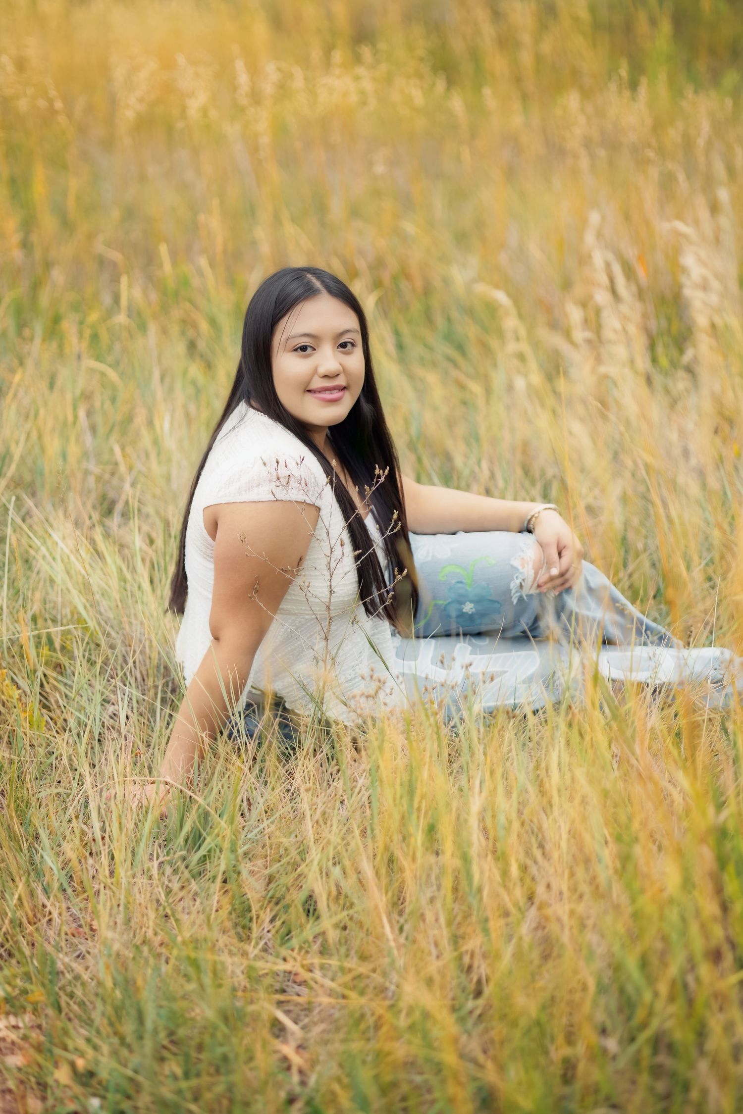 Young woman with long dark hair sits smiling in tall golden grass field, wearing white top and light blue jeans.