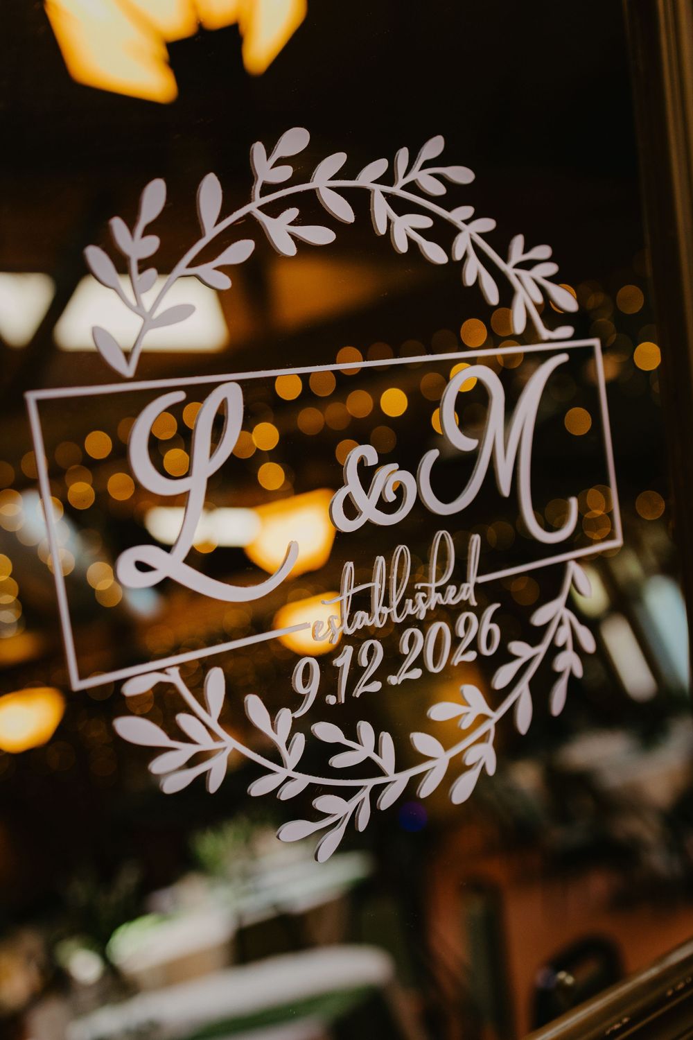 Elegant wedding monogram sign with L&M initials, floral wreath, and established date 9.12.2026 on glass.