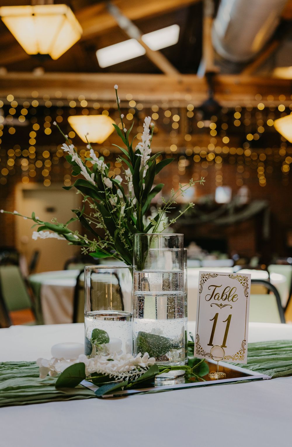 Elegant wedding reception centerpiece with white flowers, greenery, candles, and a Table 11 sign on a mirrored tray.