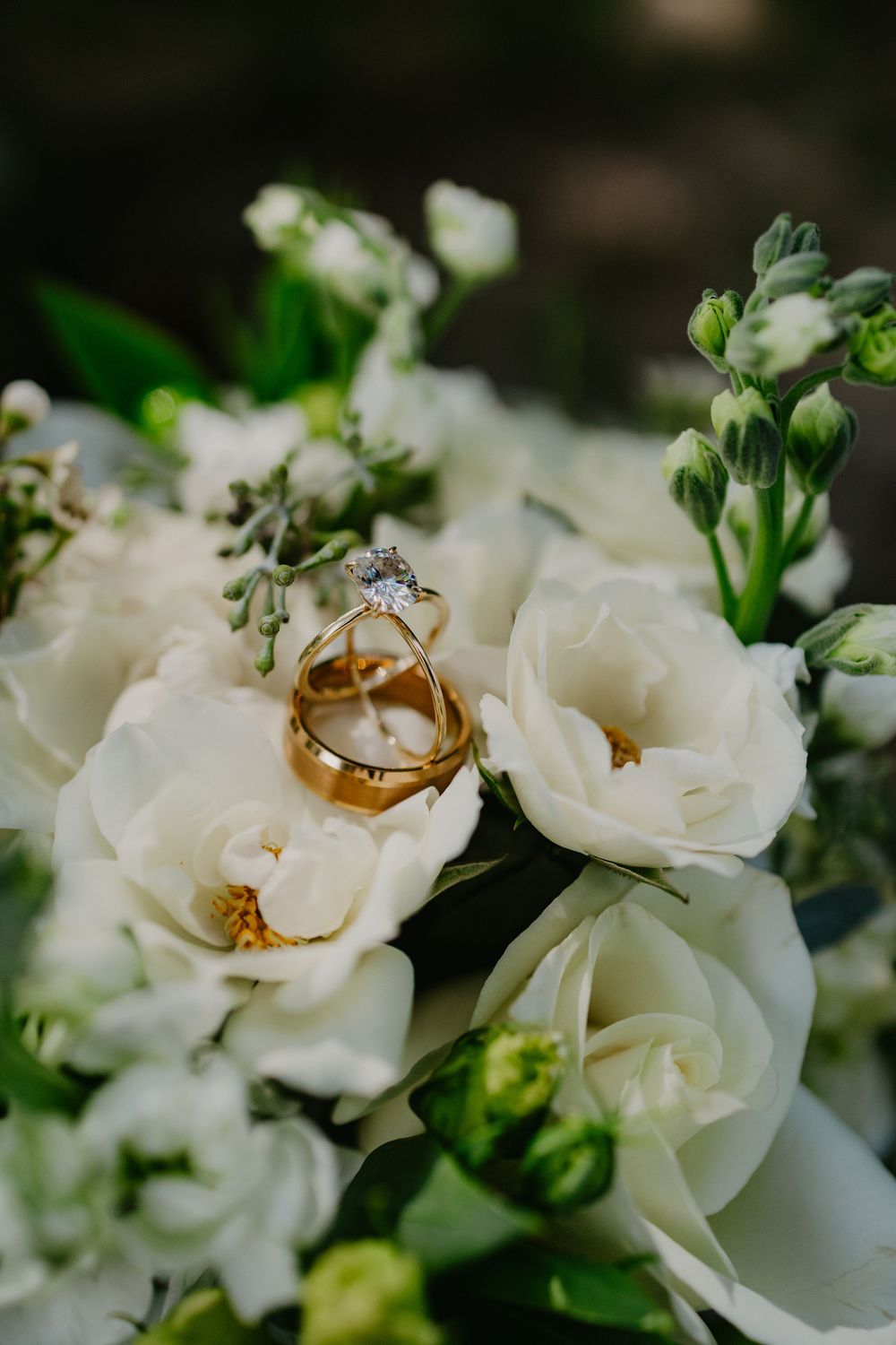 Gold wedding bands and diamond engagement ring rest on white roses and lisianthus blooms in an elegant bridal bouquet.