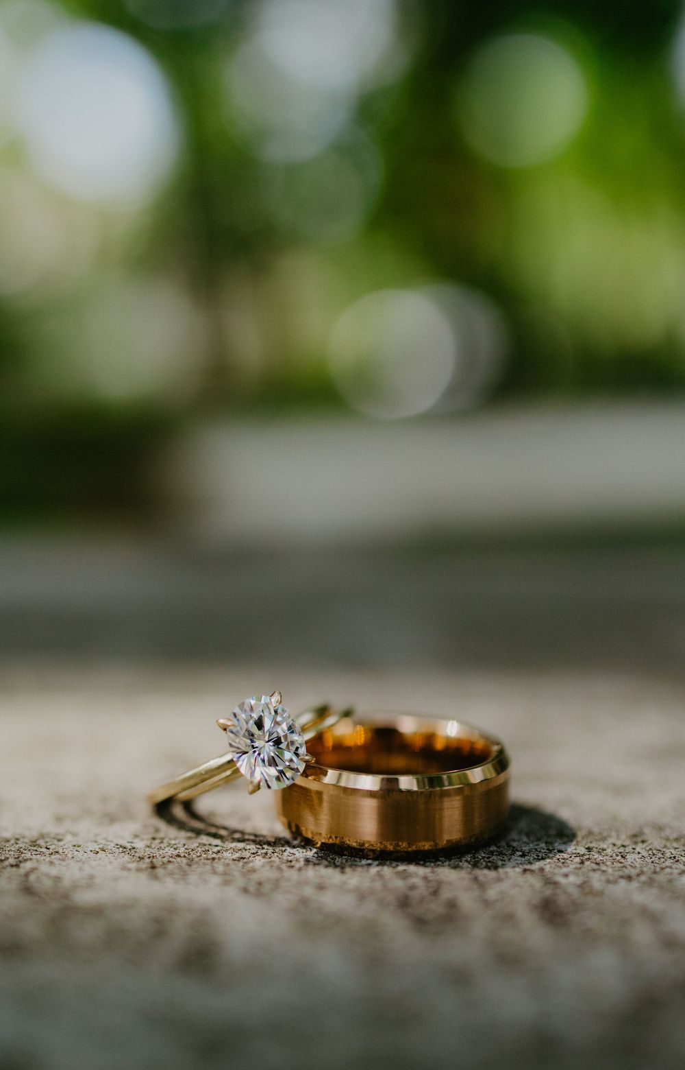 Gold wedding band and diamond solitaire engagement ring resting on stone surface with green bokeh background.