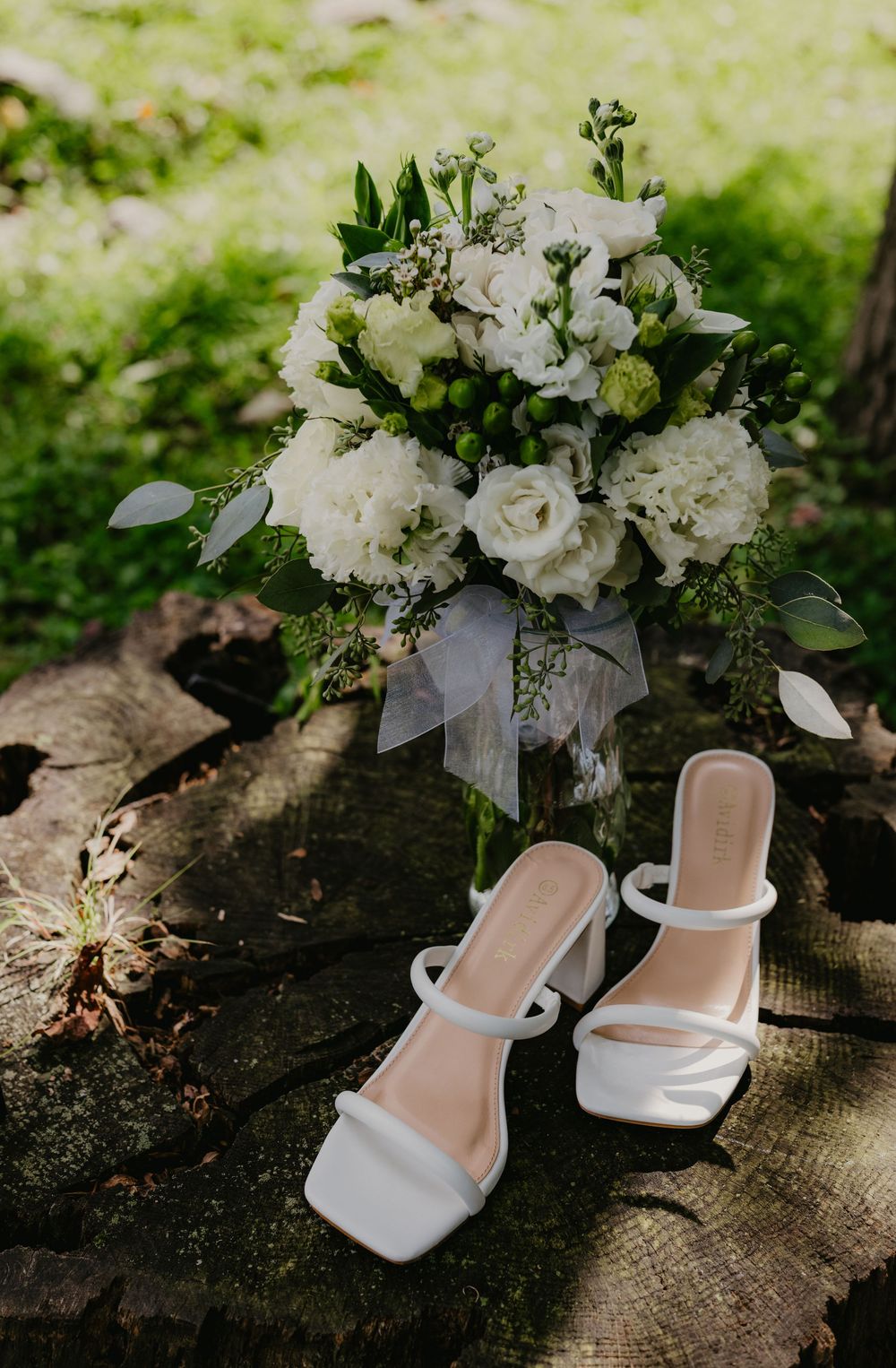 White bridal heels and a lush white floral bouquet with greenery displayed on a rustic wood stump outdoors.