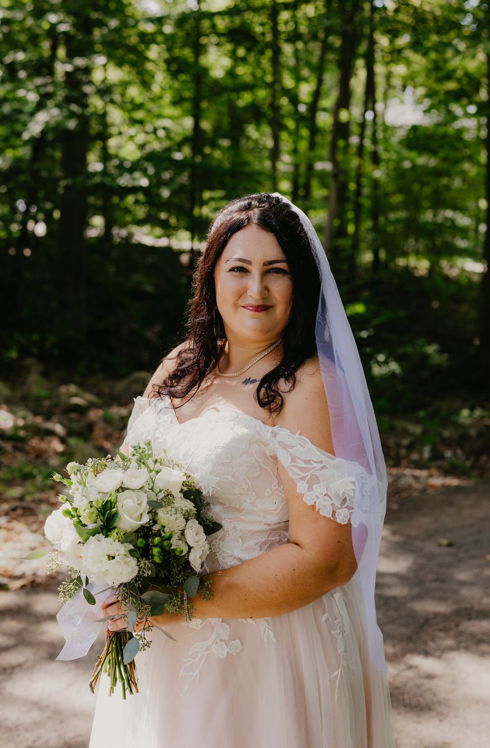 Bride in off-shoulder lace wedding gown and veil holds white bouquet, smiling outdoors in a lush green forest setting.