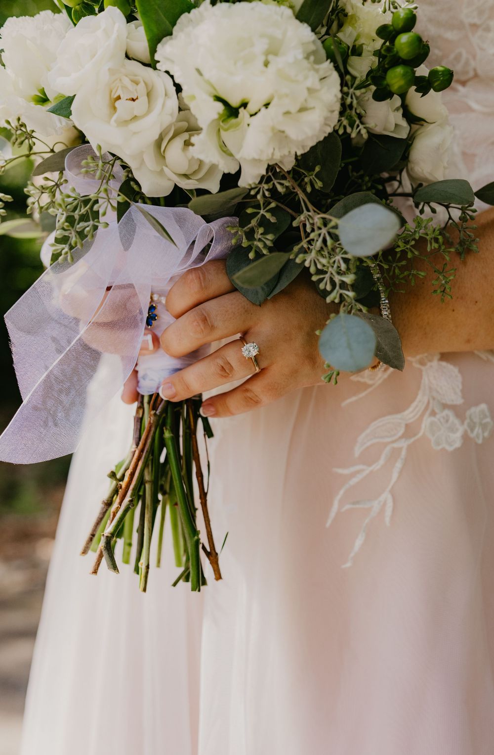 Bride holding a white floral wedding bouquet with eucalyptus and lavender ribbon, showcasing an engagement ring on her finger.