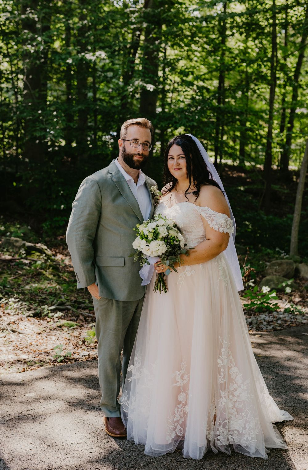 A couple poses in a forest setting, the groom in a gray suit and the bride in a white lace off-shoulder gown holding a bouquet.