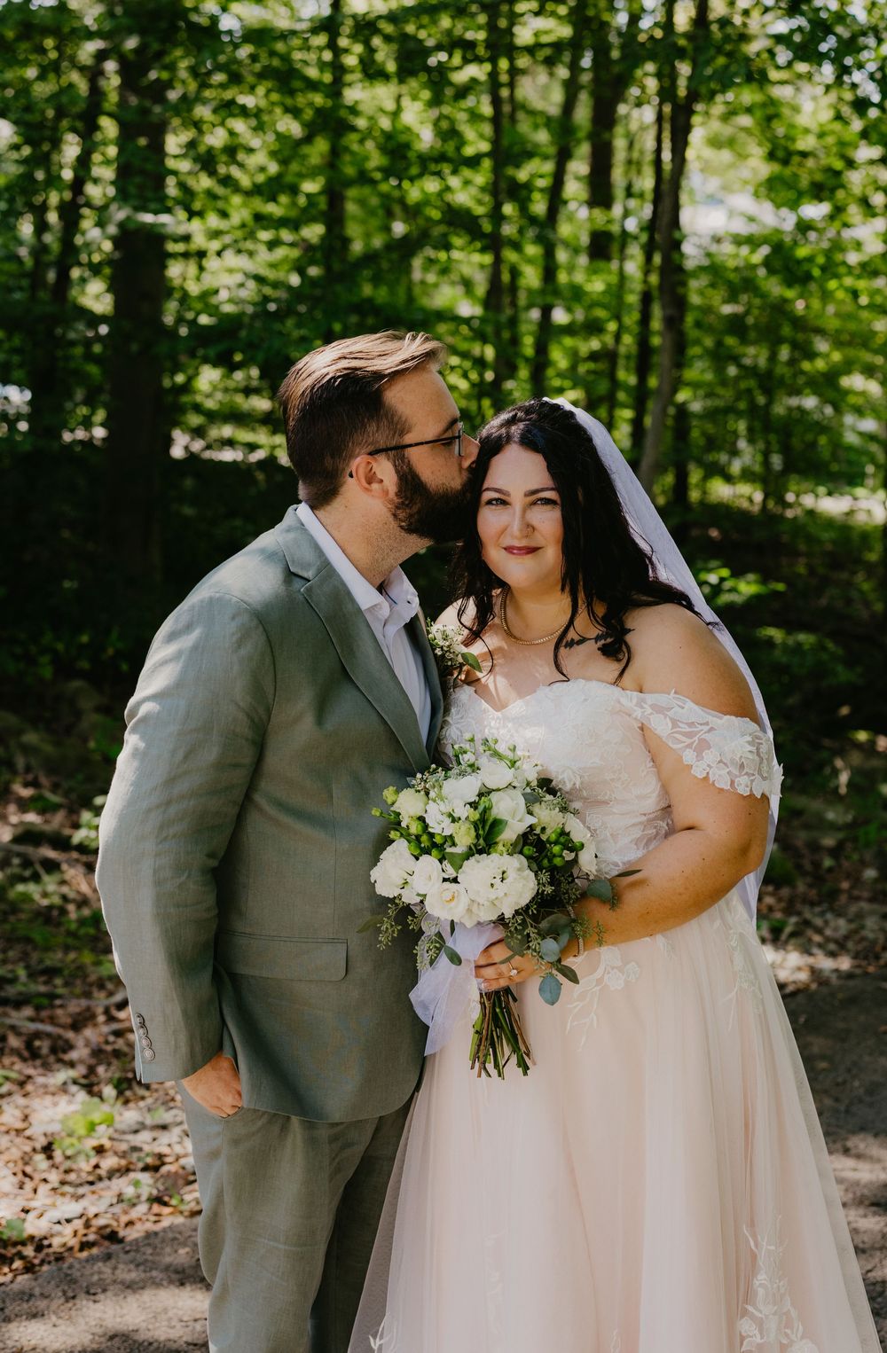 Bride in off-shoulder gown holding white bouquet stands with groom in gray suit sharing a tender moment in a sunlit forest.