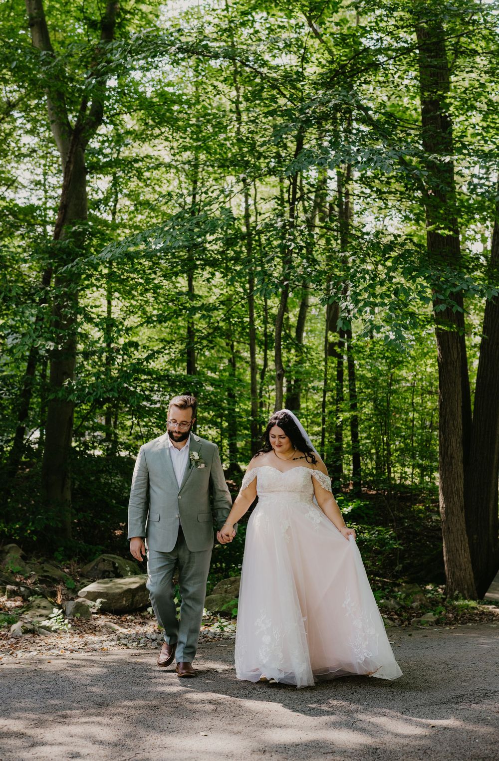 Bride in blush gown and groom in gray suit hold hands walking through lush green forest on their wedding day.