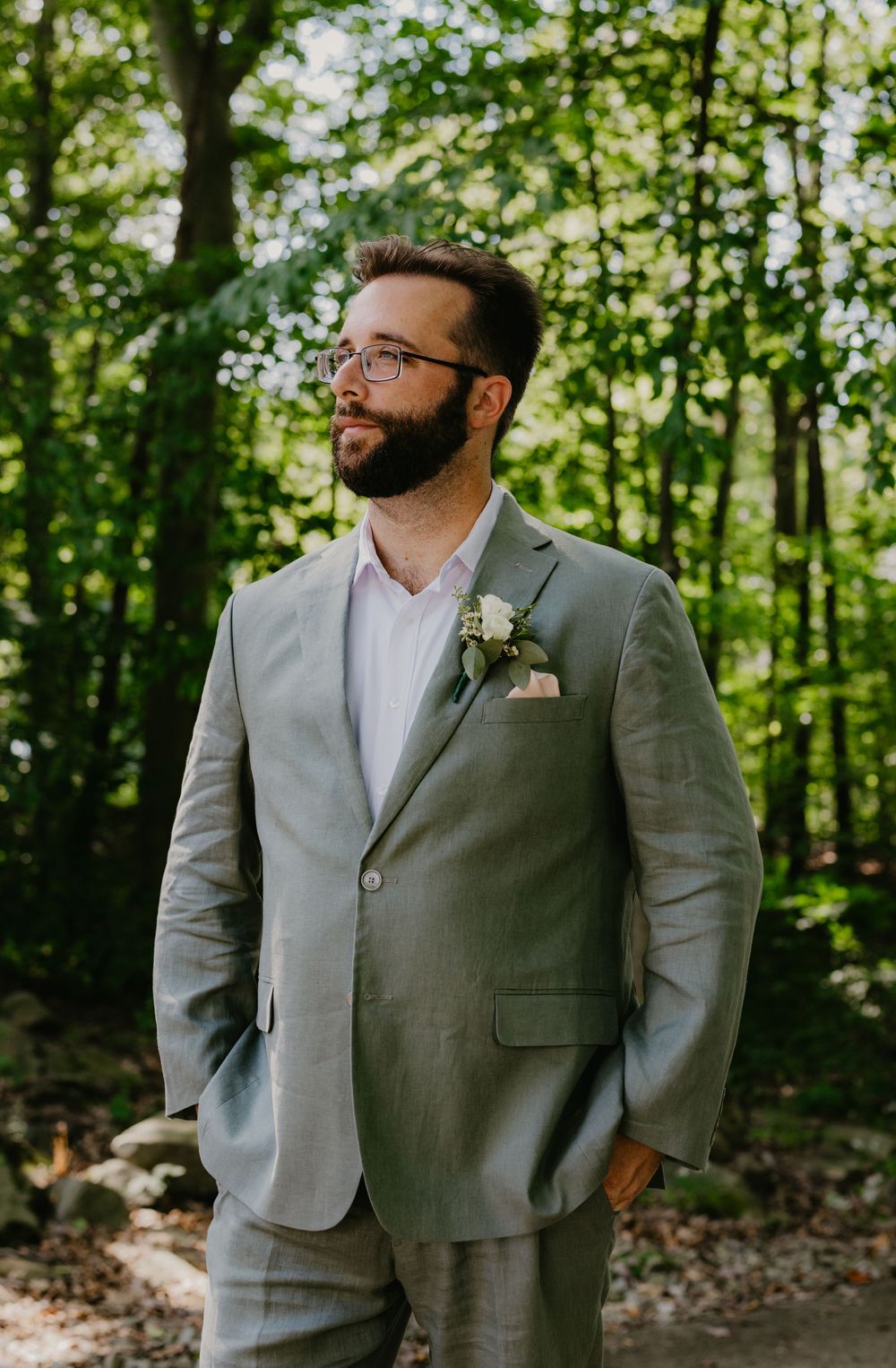 A bearded groom in a sage green suit with boutonniere and glasses stands in a lush forest setting.