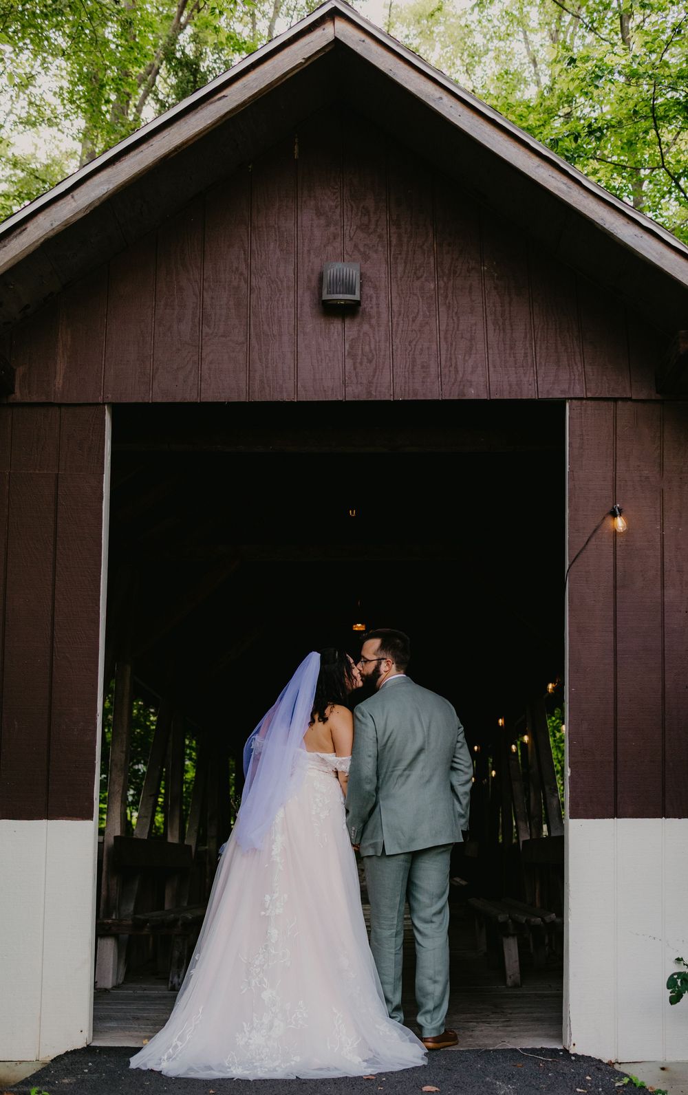 Bride and groom share a kiss at the entrance of a rustic barn wedding venue surrounded by lush green trees.