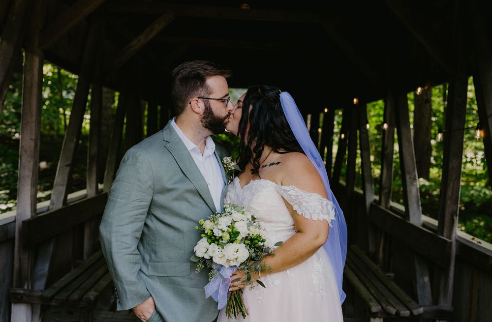 Bride and groom share a romantic kiss on a rustic covered wooden bridge surrounded by lush green trees.