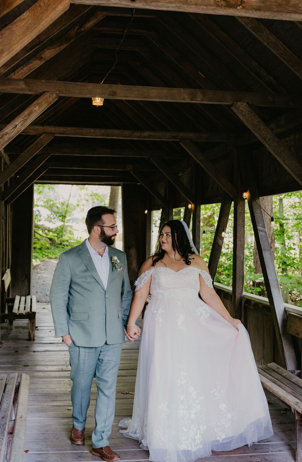 Bride and groom hold hands walking through a rustic covered bridge on their wedding day.