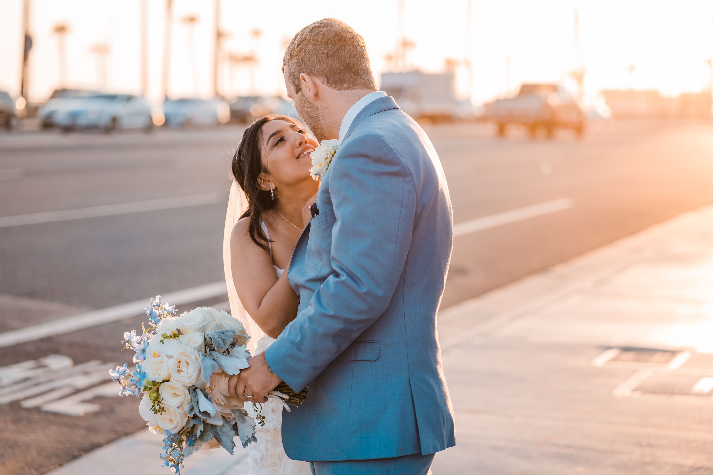 Bride and groom share a romantic moment at sunset on a coastal street, bride holding a blue and white bouquet.