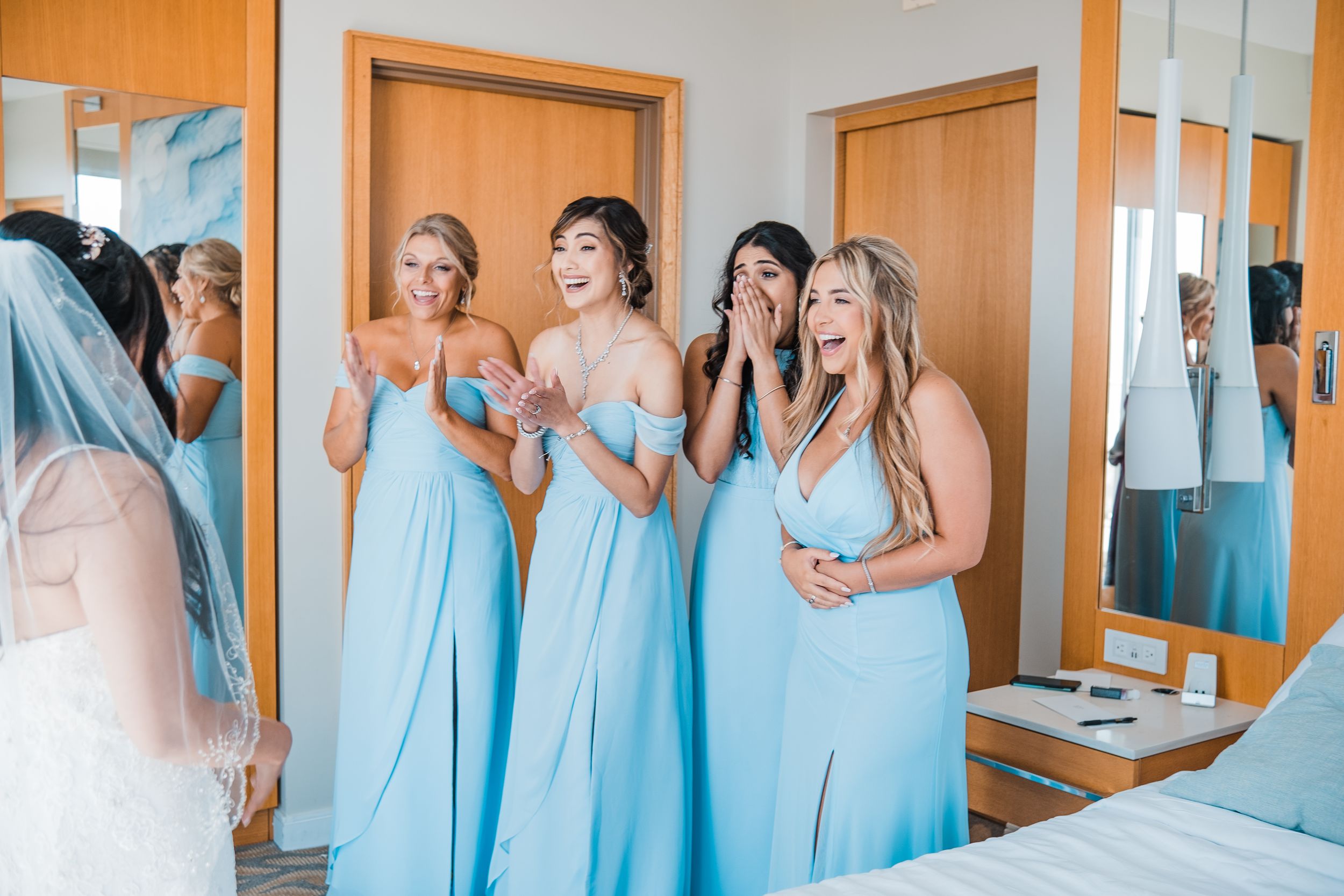 Bridesmaids in light blue gowns react with emotional surprise seeing bride in wedding dress for first time.