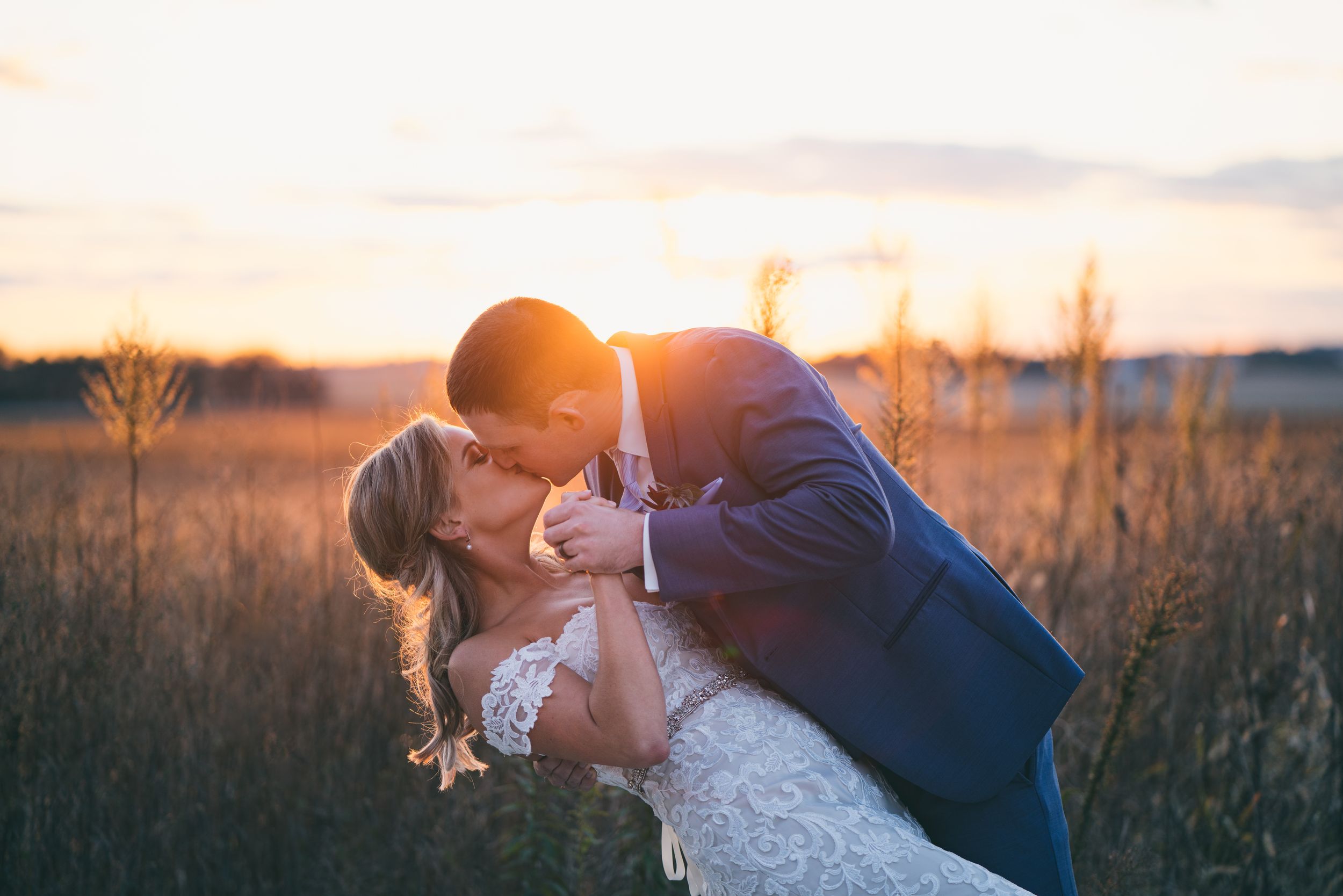 Bride and groom share a passionate dip kiss in a golden field at sunset.